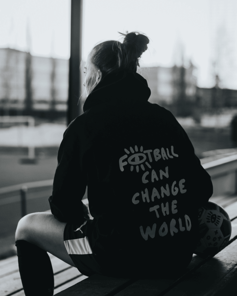A female football player sitting on a bench overlooking a football field wearing a shirt with the text "football can change the world" written on the back