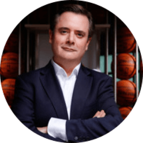 Middle aged, serious looking man in a suit standing with his arms corssed in front off basketballs, he's looking into the camera